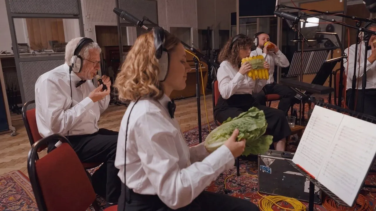 Musicians in formal shirts wear headphones and eat vegetables in a recording studio.