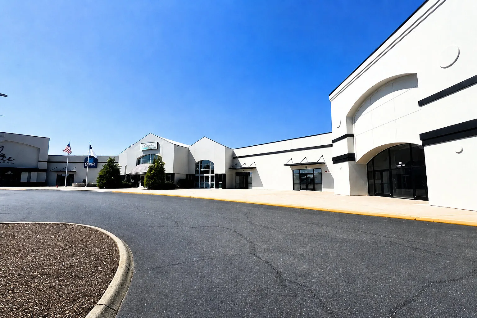 Exterior of a shopping center with storefronts, driveway and clear blue sky.