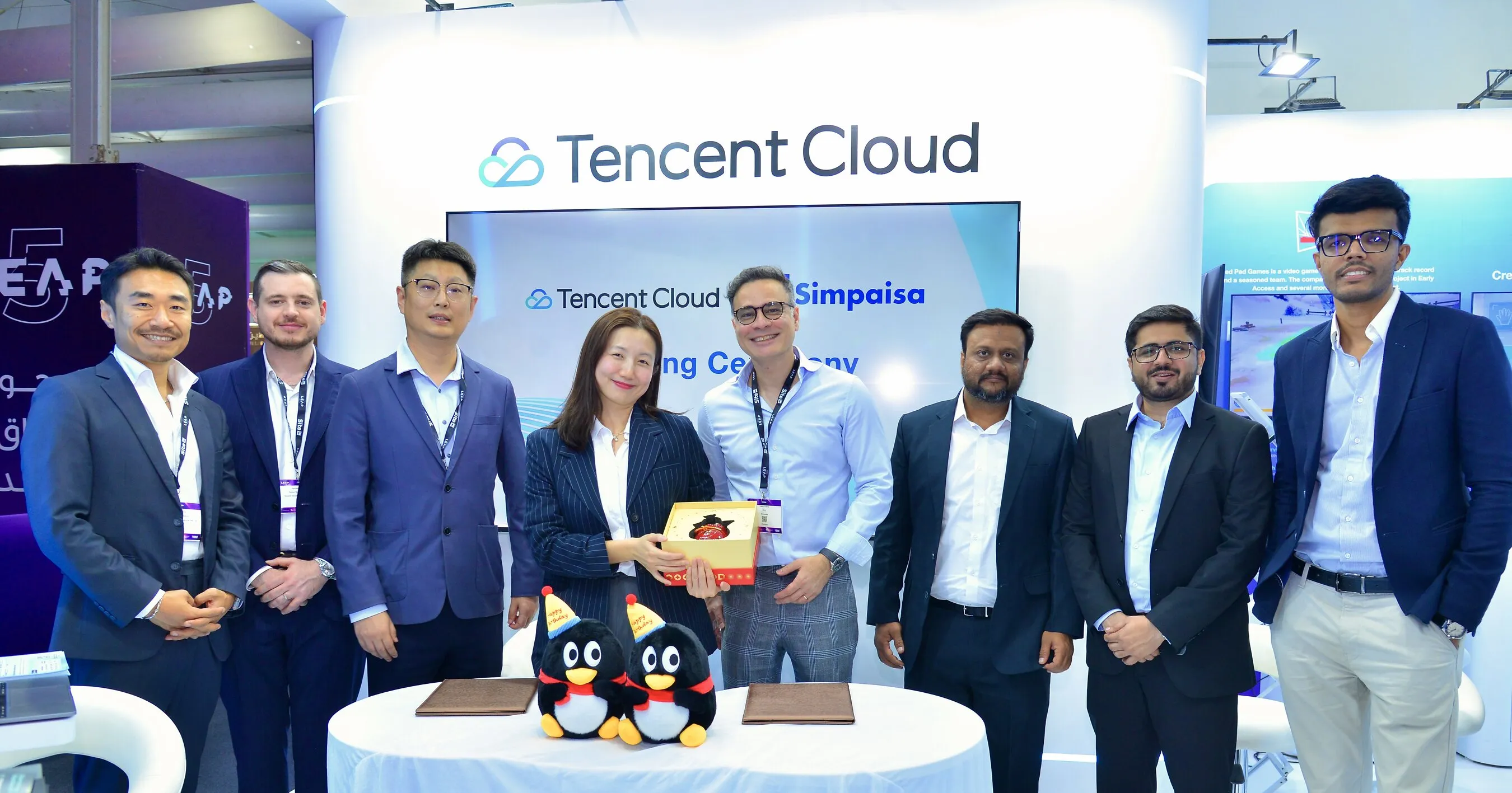 Eight people at a Tencent Cloud booth, with plush penguins and a gift box on a table.