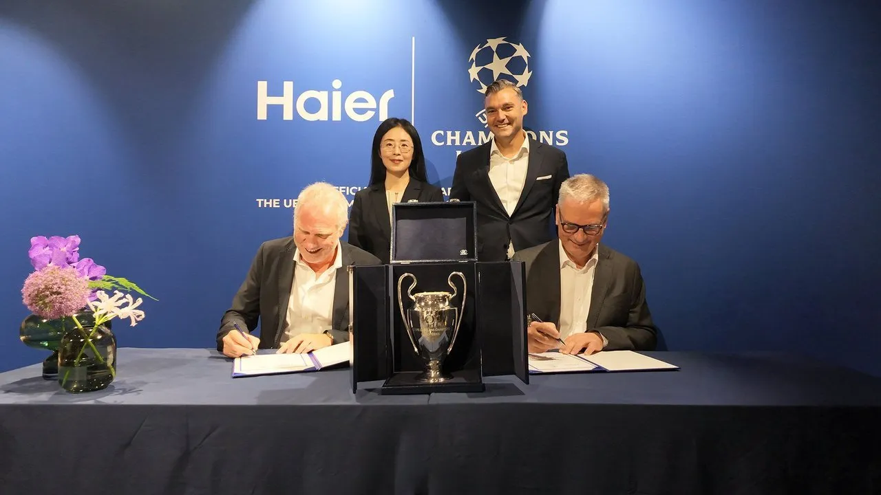 Two men sign documents beside a UEFA Champions League trophy as two people stand behind them.