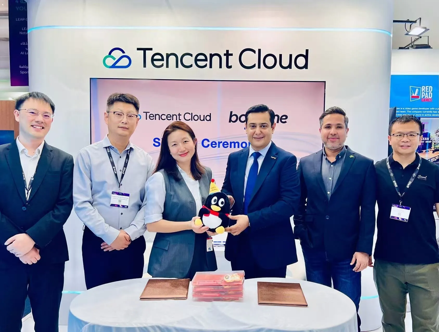 Six people at a Tencent Cloud booth hold a penguin plush toy during a signing ceremony.
