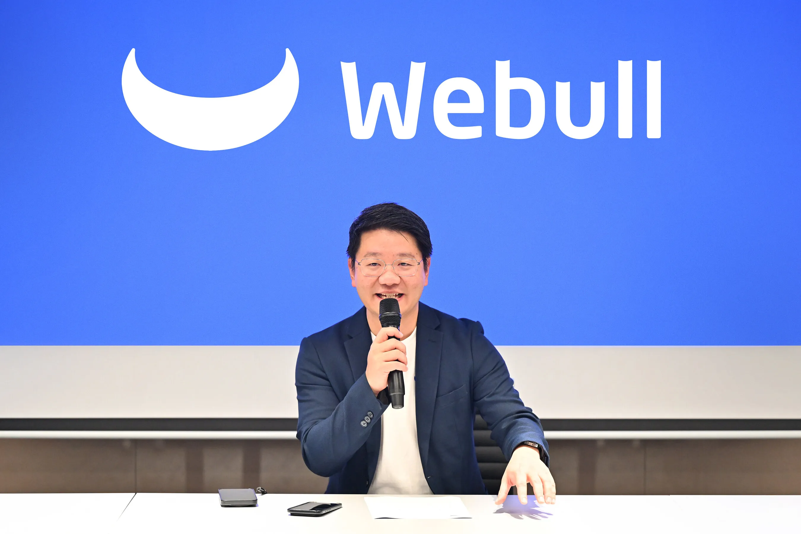 A man speaks into a microphone in front of a blue Webull logo backdrop.