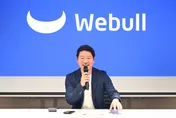 A man speaks into a microphone in front of a blue Webull logo backdrop.
