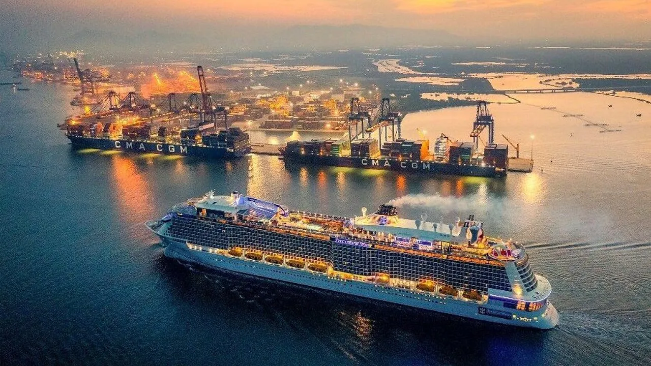 A brightly lit cruise ship sails past container ships and cranes at a harbor at sunset.