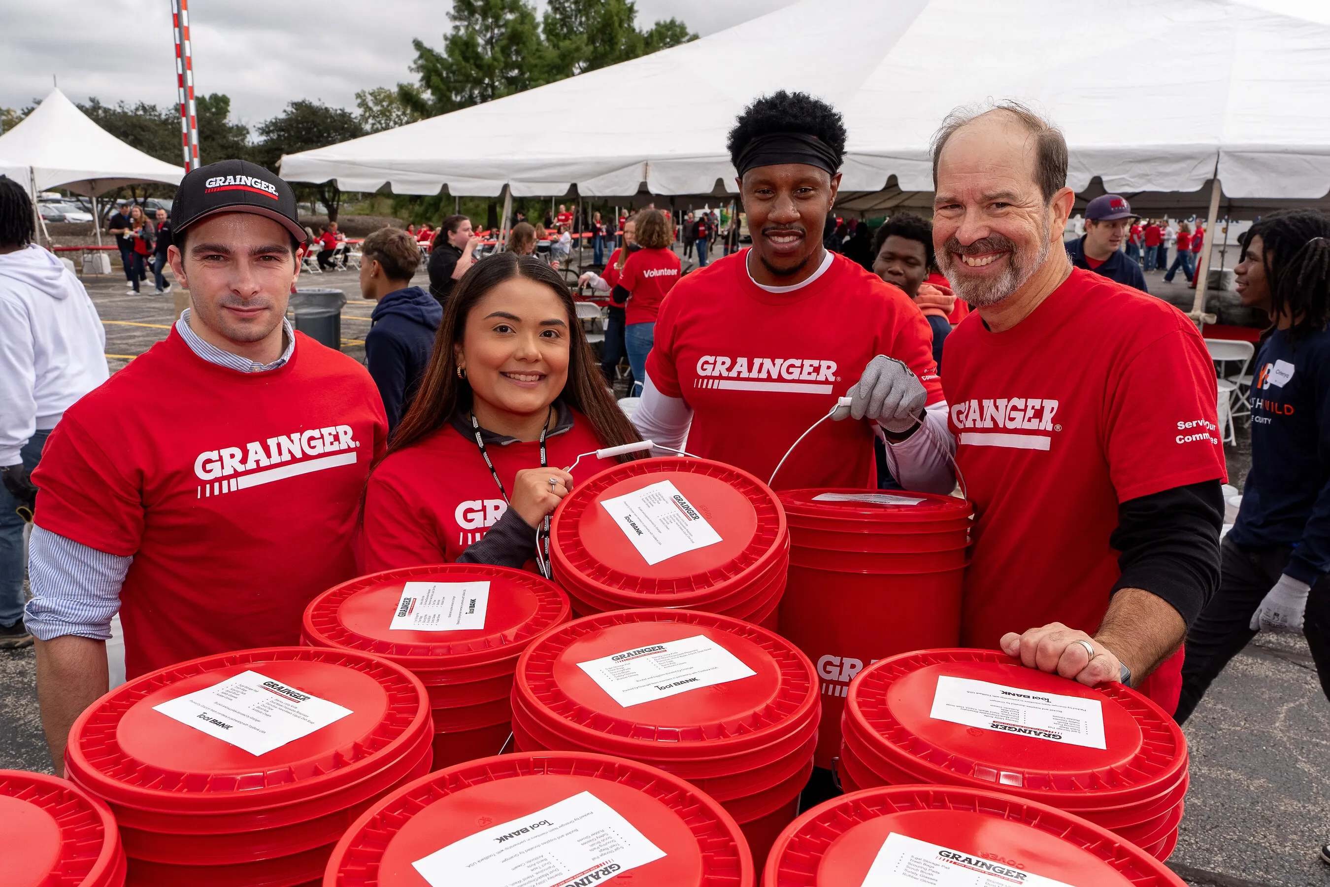 Four volunteers in red Grainger shirts stand behind red buckets under a tent.