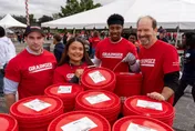 Four volunteers in red Grainger shirts stand behind red buckets under a tent.