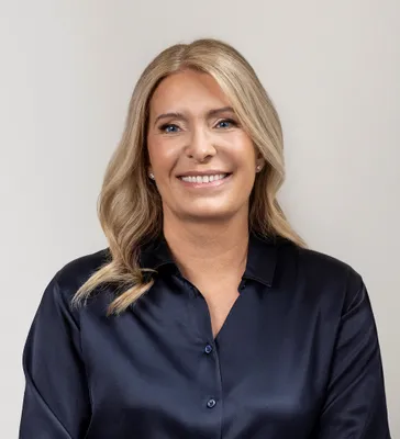 Carolina Dybeck Happe, smiling, in a dark blue blouse against a plain light grey background.