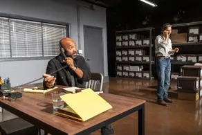Two men talk on phones in an office filled with shelves of labelled boxes.