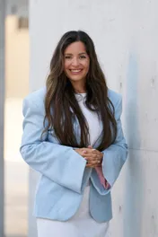 Smiling woman in a light blue blazer standing against a white wall.