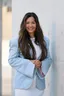 Smiling woman in a light blue blazer standing against a white wall.