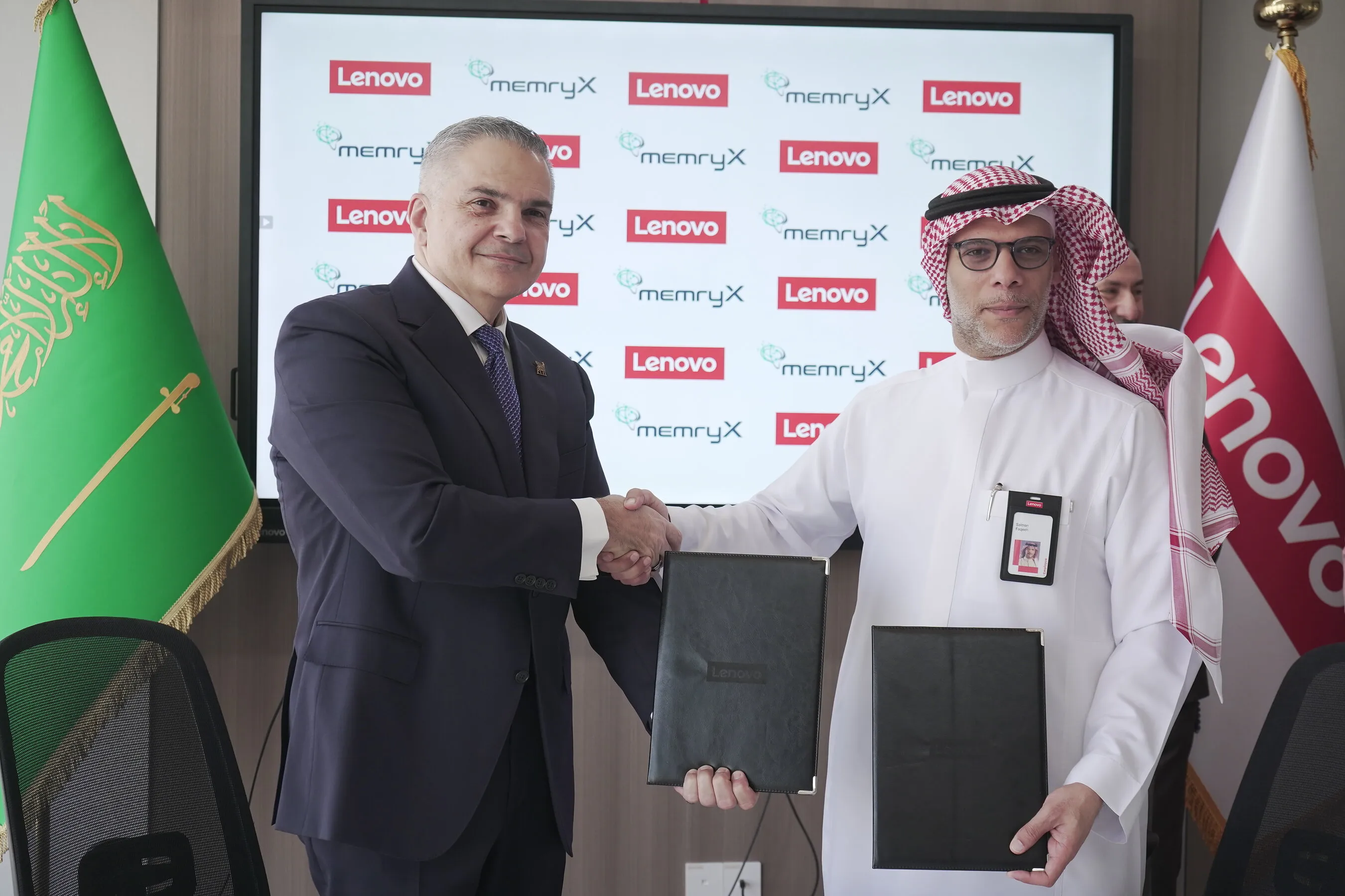 Two men shake hands while holding document folders before Lenovo and memryX logos and Saudi Arabian flags.