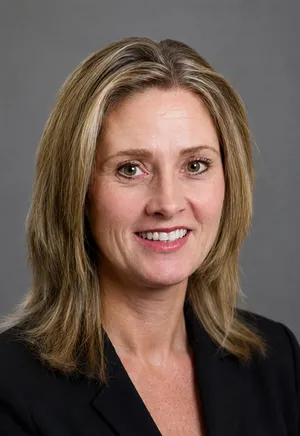 Head-and-shoulders portrait of a smiling woman with blonde hair wearing a black blazer against a gray background