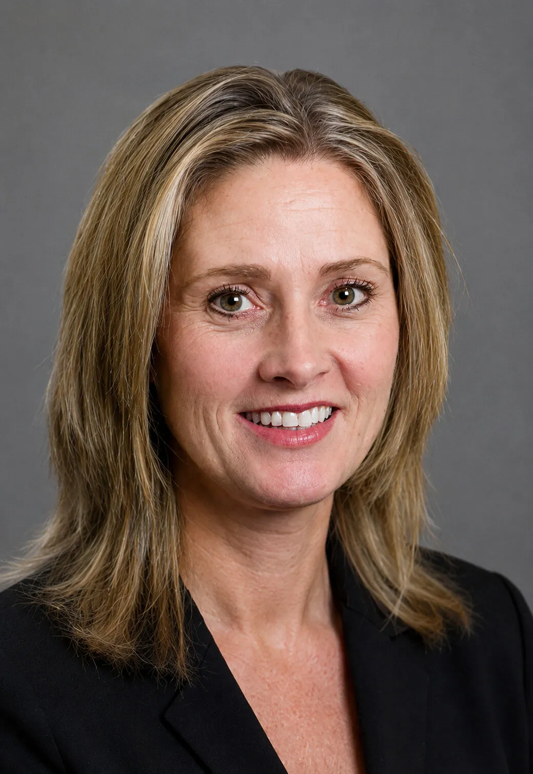 Head-and-shoulders portrait of a smiling woman with blonde hair wearing a black blazer against a gray background
