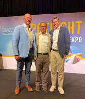 Three men in business-casual clothing stand on a stage before an event backdrop.