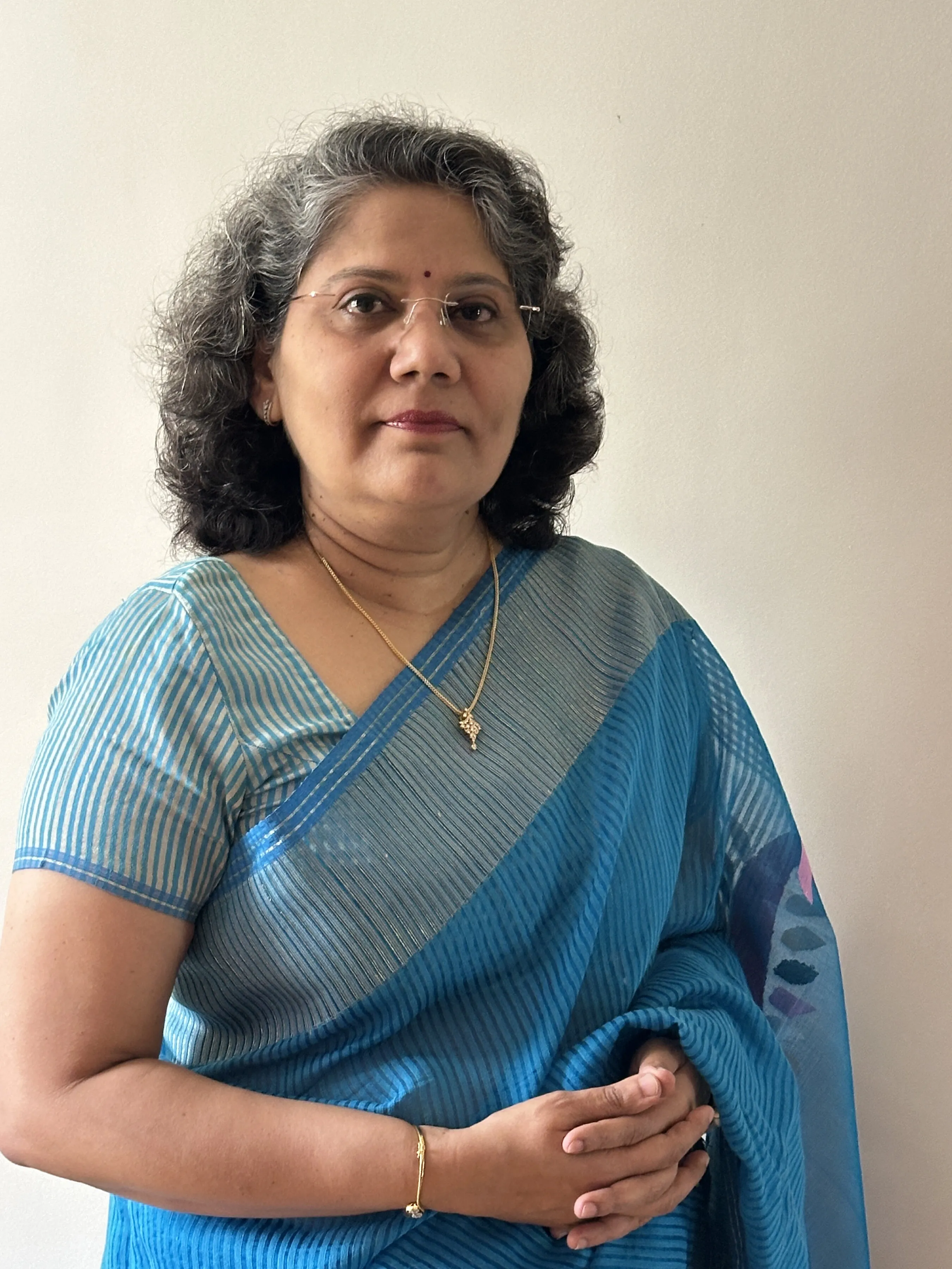 Jyotsna Kher wearing a blue sari, standing with her hands clasped.