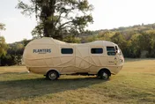 A peanut-shaped Planters camper van parked on a grassy field beneath trees