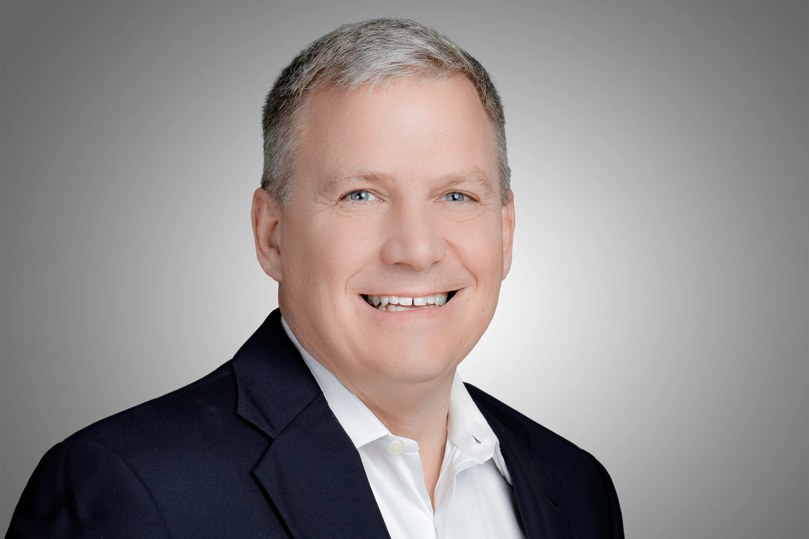Richard Tudgay smiling in a dark suit jacket and white shirt against a grey background.