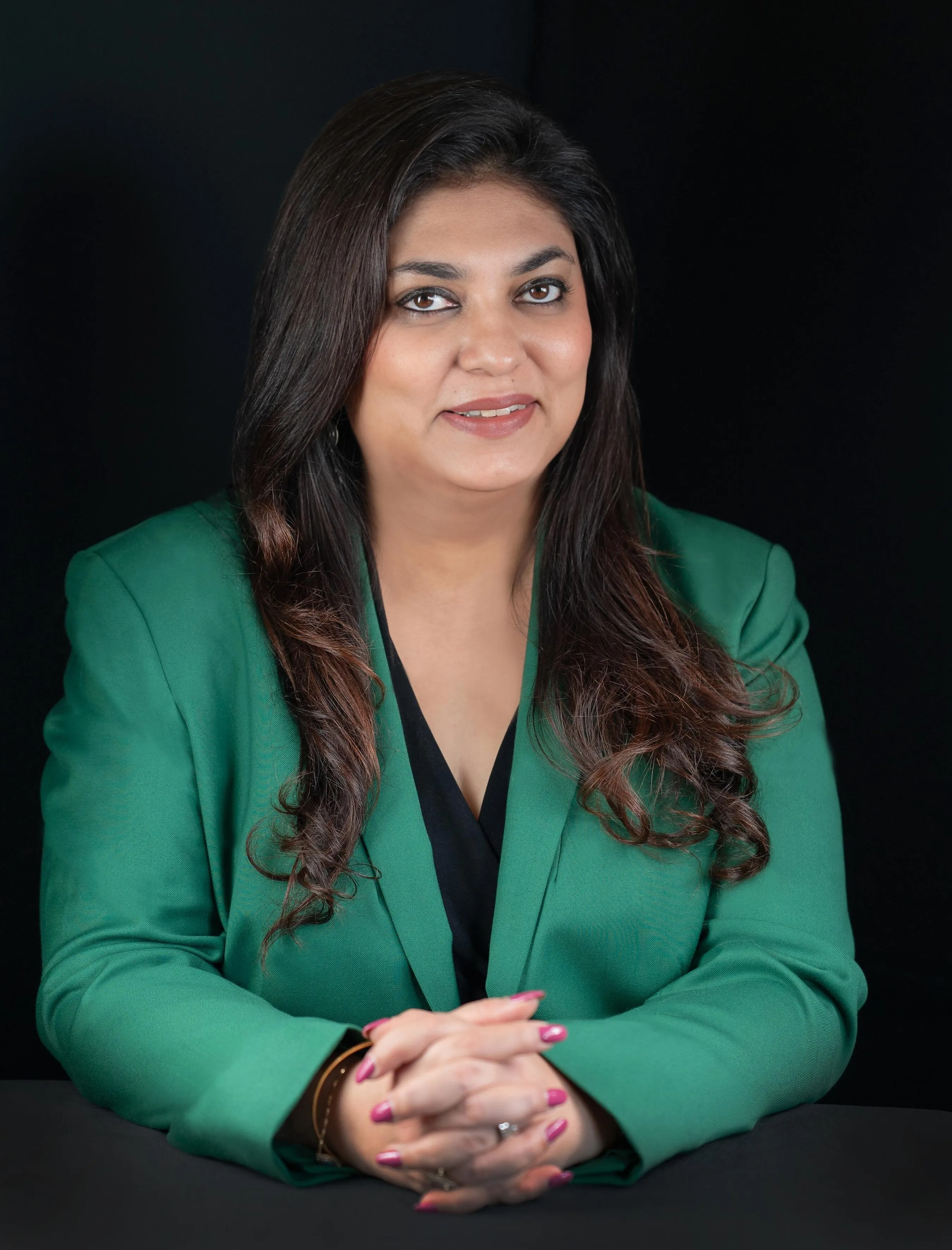 Shweta Rajpal Kohli in a green blazer, seated with hands clasped against a black background.