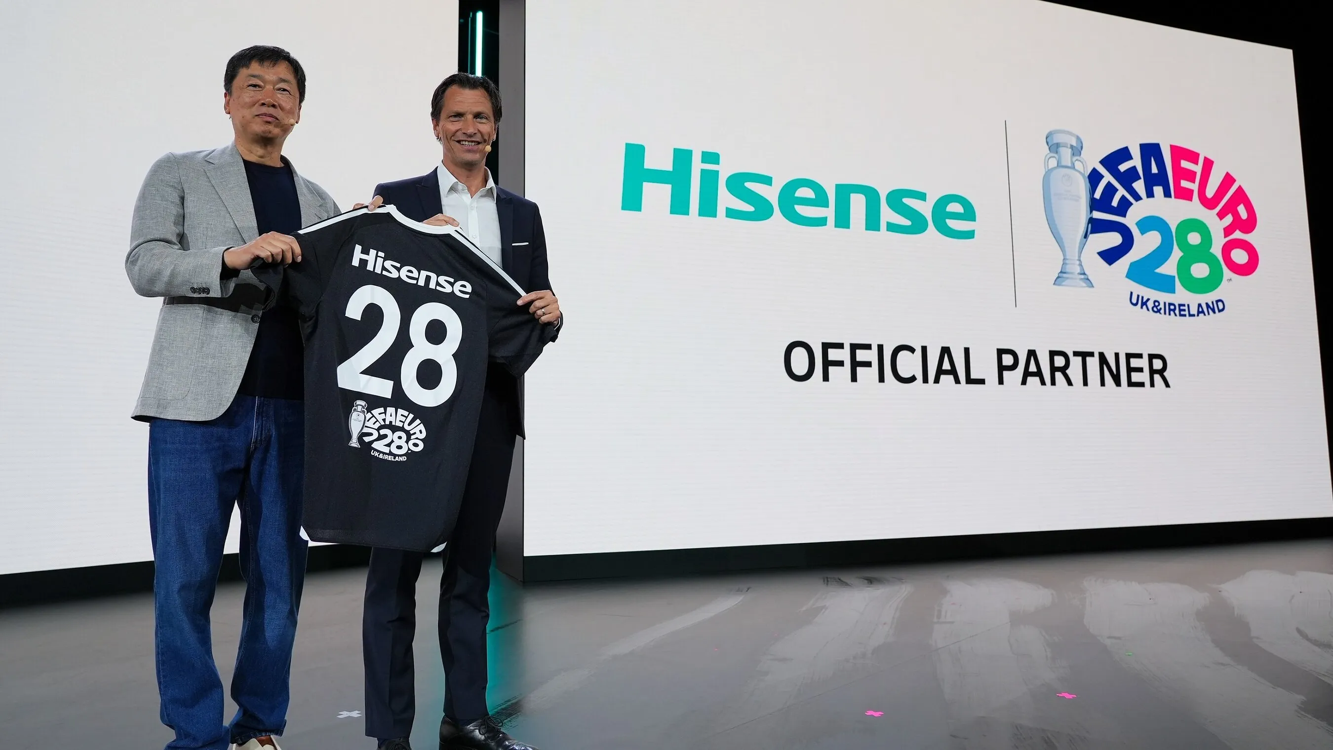 Two men hold a Hisense number 28 football shirt beside a UEFA Euro 2028 official partner display.