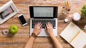 Hands typing on a laptop beside a phone, notebook, pencils, coffee and desk items.