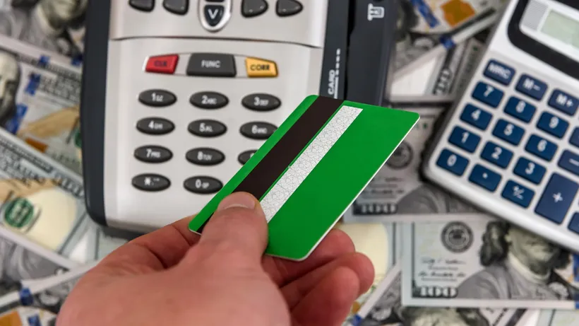 Hand holding a green card over calculators, cash, and pens.