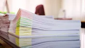 Stacks of paperwork with coloured folders on a desk in a bright office.