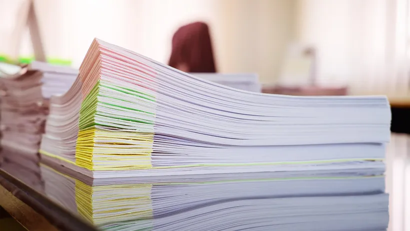 Stacks of paperwork with coloured folders on a desk in a bright office.