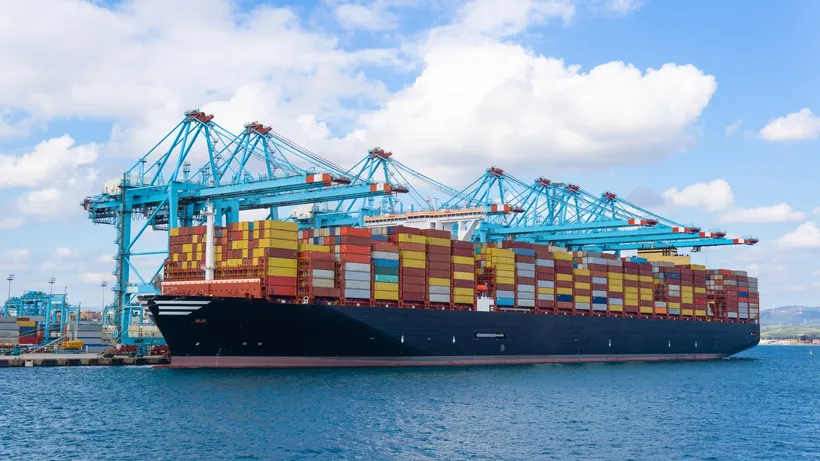 Container ship loaded with colourful containers docked beside large blue cargo cranes under a cloudy sky.