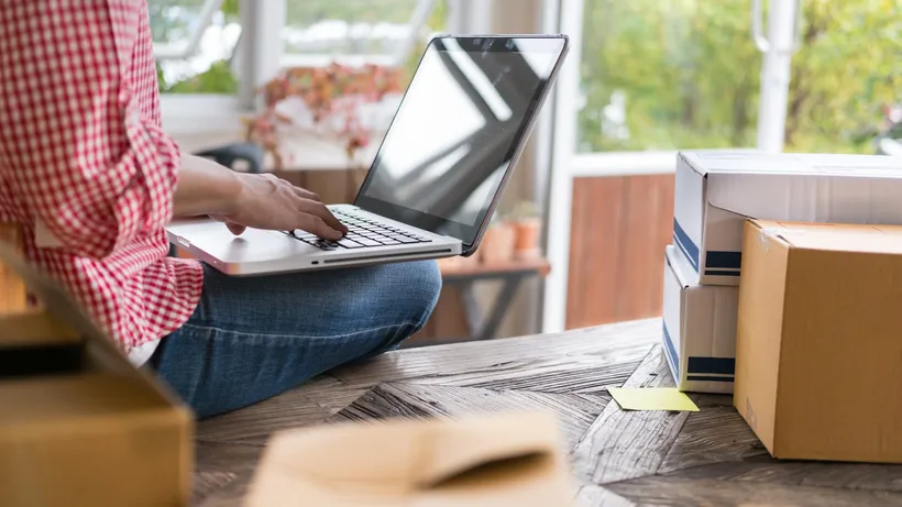 Person using a laptop beside stacked cardboard boxes in a bright room.
