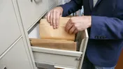 Person in a blue jacket sorting paper files in an open filing cabinet.