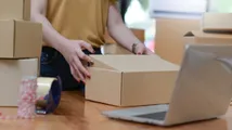 Person sealing a cardboard box beside packing tape, stacked boxes, and a laptop.