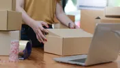 Person sealing a cardboard box beside packing tape, stacked boxes, and a laptop.