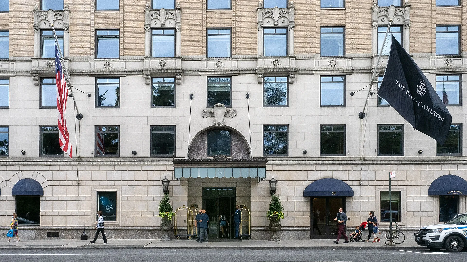 The entrance of The Ritz-Carlton New York, Central Park, with hotel staff at the door, luggage trolleys and a Ritz-Carlton flag