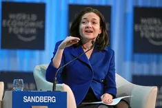 Sheryl Sandberg in a blue jacket, seated in a white armchair, speaking into a microphone behind a name card reading Sandberg.
