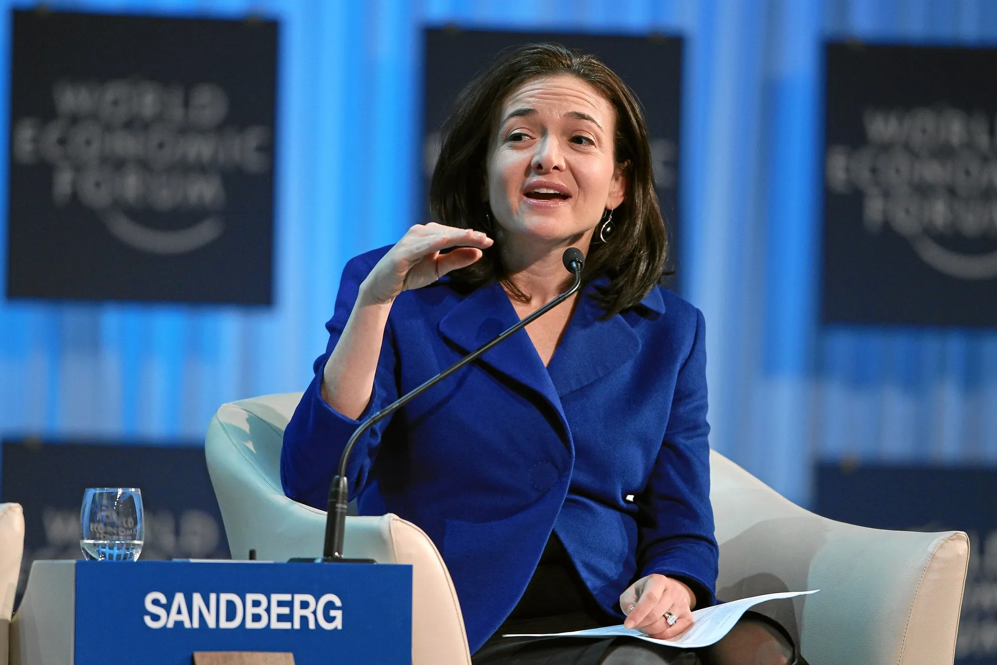 Sheryl Sandberg in a blue jacket, seated in a white armchair, speaking into a microphone behind a name card reading Sandberg.